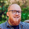 A man with a ginger beard and black glasses smiling at the camera. He is wearing a casual button down shirt, and there is green foliage in the background.